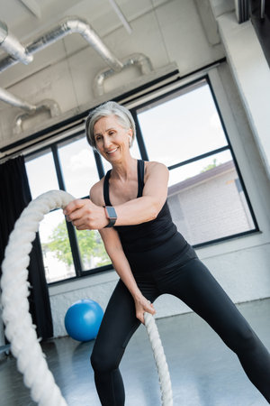 cheerful senior sportswoman with grey hair working out with battle ropes in gym