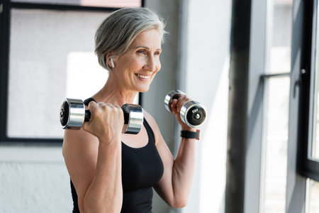 happy and senior woman in wireless earphone exercising with dumbbells in gymの写真素材