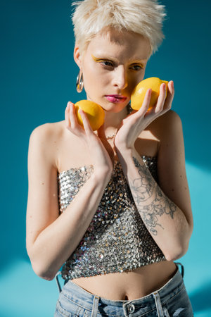 tattooed albino woman with trendy makeup and blonde hair posing with ripe lemons on blue