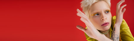 tattooed albino woman with yellow eyeliner looking away while posing on carmine pink, bannerの写真素材