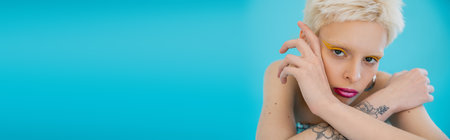 tattooed and blonde albino model with bright makeup looking at camera on blue background, bannerの写真素材