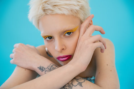 tattooed and blonde albino model with bright makeup looking at camera on blue backgroundの写真素材