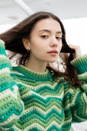 Portrait of young woman in sweater touching hair and looking at camera during trip on yacht