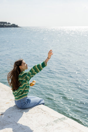 Side view of young woman holding bread while sitting on pier in Turkeyの写真素材