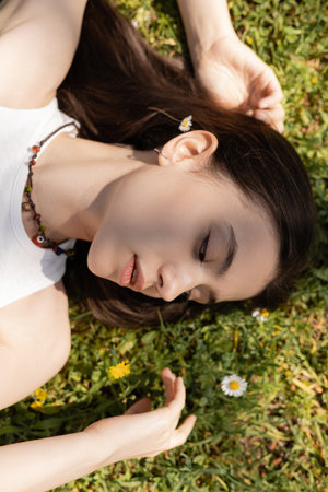 Top view of young brunette woman looking at flowers while lying on lawn in parkの写真素材