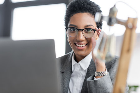 portrait of cheerful african american businesswoman adjusting stylish eyeglasses and looking at camera near blurred laptop in officeの写真素材
