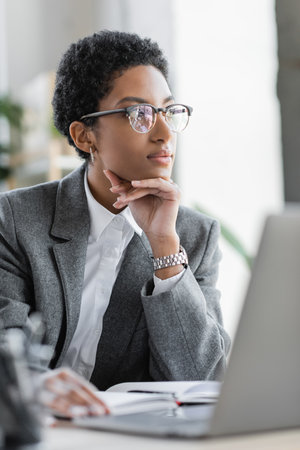 thoughtful african american businesswoman in grey blazer and eyeglasses looking away in officeの写真素材