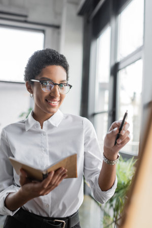 positive african american businesswoman in stylish eyeglasses holding notebook and pointing with pen in officeの写真素材