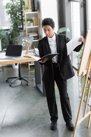 full length of african american businesswoman in black suit standing with documents near note board in contemporary officeの写真素材