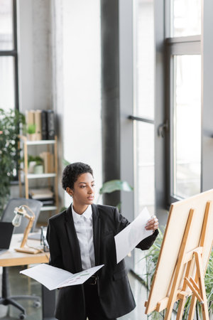 young african american businesswoman holding papers and looking at note board while working in officeの写真素材