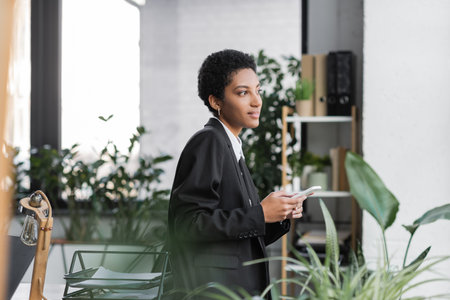 side view of smiling african american businesswoman in black blazer holding smartphone and looking away in officeの写真素材