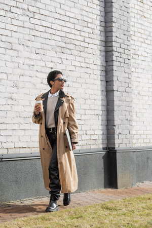 full length of african american businesswoman in sunglasses and trench coat walking with laptop and coffee to go near grey wall on urban streetの写真素材