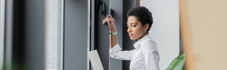 side view of young african american businesswoman in white blouse holding laptop near windows in office, bannerの写真素材