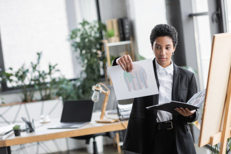 young african american businesswoman holding folder and graphs near note board in modern officeの写真素材