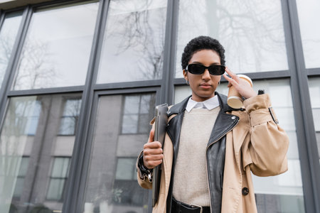 stylish african american businesswoman with paper cup and laptop adjusting sunglasses near urban building with glass facadeの写真素材