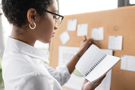 side view of african american businesswoman standing near blurred note board and holding blank notebook in officeの写真素材