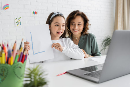 Smiling woman looking at cute daughter with letter on paper having speech therapy video call at homeの写真素材