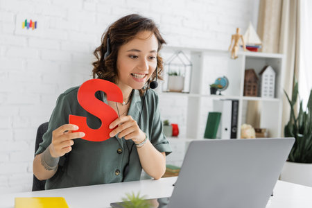 Smiling speech therapist in headset holding letter s during video call on laptop in consulting room