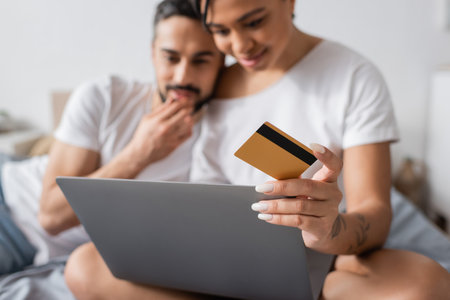 selective focus of credit card and laptop near blurred interracial couple thinking during online shopping in bedroom at homeの写真素材