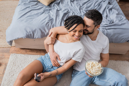 high angle view of cheerful man with bowl of popcorn embracing african american woman with tv remote controller on floor in bedroomの写真素材
