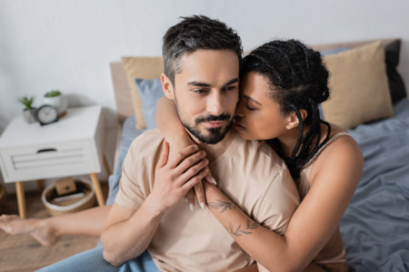 tattooed african american woman embracing bearded man in t-shirt in bedroom at homeの写真素材