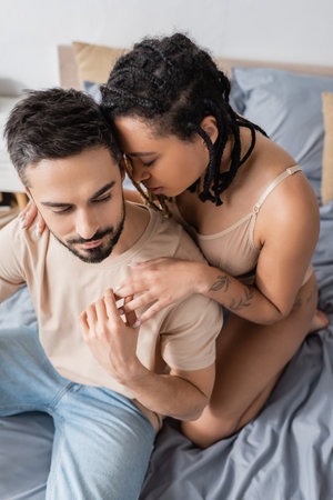 high angle view of bearded man touching hand of hot african american woman in lingerie in bedroom at homeの写真素材
