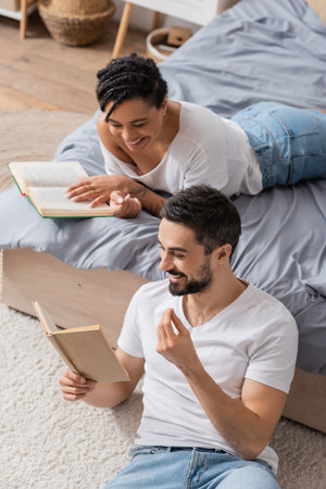 high angle view of cheerful bearded man reading book to smiling african american woman lying on bed at homeの写真素材