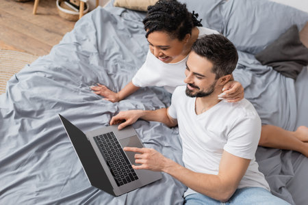 high angle view of cheerful bearded man and african american woman watching movie on computer on bed at homeの写真素材