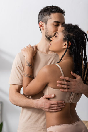 brunette man in t-shirt embracing tattooed african american woman in sexy underwear in bedroom at homeの写真素材