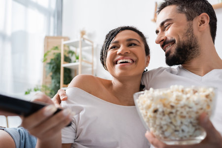 smiling bearded man with bowl of popcorn hugging carefree african american woman clicking tv channels in bedroom at homeの写真素材