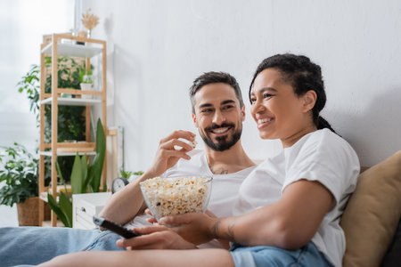 pleased bearded man eating popcorn and looking at smiling african american woman with tv remote controller on bed at homeの写真素材