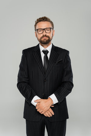 confident and bearded businessman in formal wear and glasses looking at camera isolated on greyの写真素材
