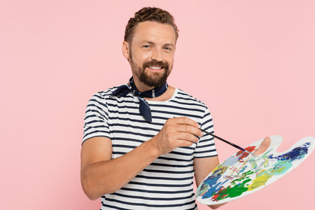 positive french artist with neck scarf holding palette with colorful paints and paintbrush isolated on pinkの写真素材
