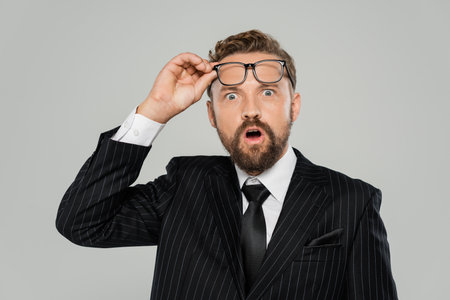 shocked businessman in formal wear adjusting glasses and looking at camera isolated on greyの写真素材