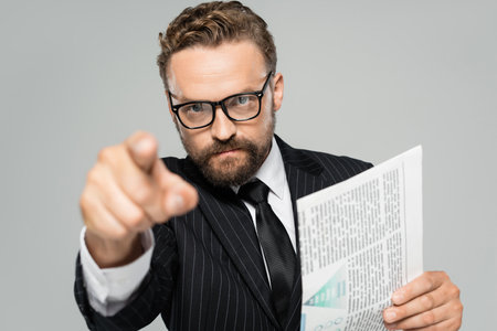 businessman in suit and glasses pointing with finger at camera while holding newspaper isolated on greyの写真素材