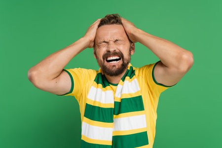 emotional sports fan in striped scarf and yellow t-shirt pulling his hair isolated on greenの写真素材