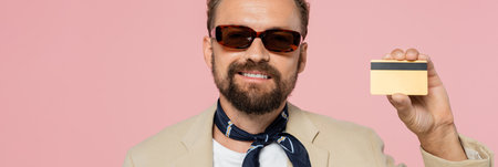 overjoyed man in stylish sunglasses and neck scarf holding credit card isolated on pink, banner