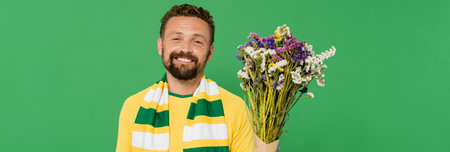 cheerful and bearded football fan holding wildflowers isolated on green, banner
