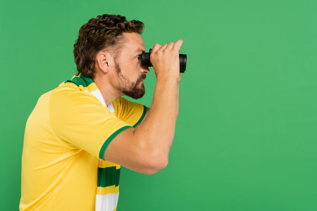 side view of bearded man in striped scarf holding binoculars during football match isolated on greenの写真素材