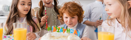 redhead boy blowing candles on birthday cake near friends during party at home, bannerの写真素材