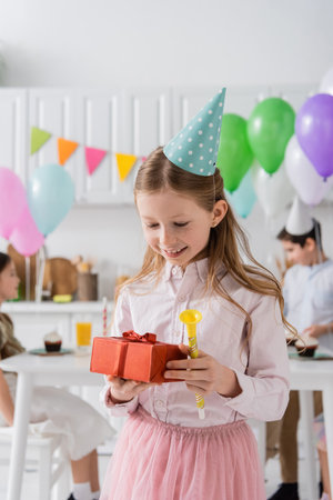 happy preteen girl holding birthday present and party horn near friends on blurred backgroundの写真素材