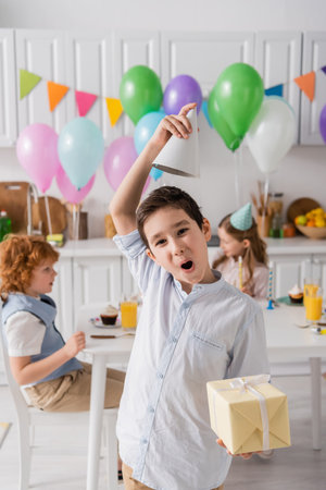 funny preteen boy in braces holding birthday present and adjusting party cap near friends on blurred backgroundの写真素材