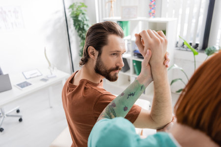 blurred physiotherapist examining arm and elbow of bearded man during appointment in rehabilitation...