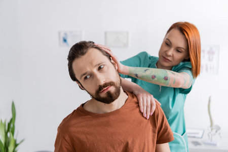 tattooed physiotherapist stretching neck of bearded man while doing diagnostics in consulting roomの写真素材