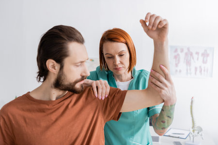 redhead physiotherapist flexing arm and touching shoulder of injured man in consulting roomの写真素材