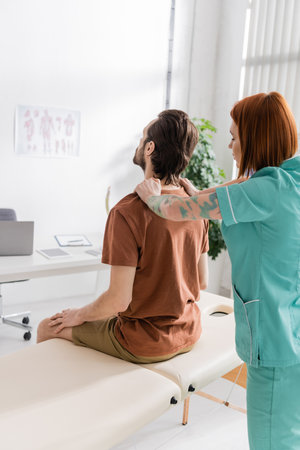 redhead osteopath touching painful back of man sitting on massage table in consulting roomの写真素材