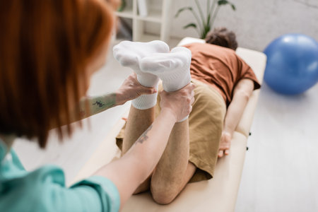 blurred physiotherapist flexing legs of injured man during recovery manipulation on massage table in...