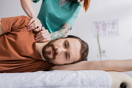 tattooed physiotherapist massaging painful shoulder of bearded man in rehab centerの写真素材