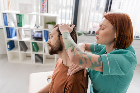 side view of tattooed physiotherapist stretching neck of bearded man during examination in rehab centerの写真素材