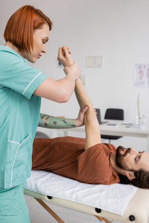 redhead physiotherapist stretching painful arm of bearded man lying on massage table in rehabilitation centerの写真素材